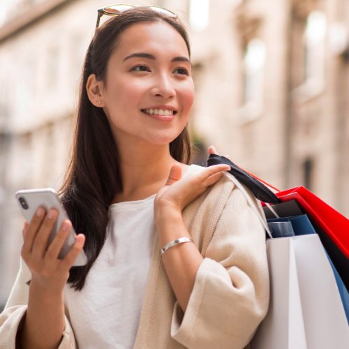 smiley-woman-holding-shopping-bags-smartphone-outdoors